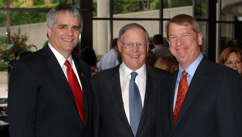 Three individuals smiling, standing side by side at an event, dressed in business attire. The person in the middle is wearing glasses.